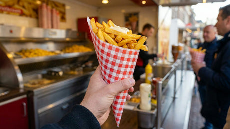 Persoon die een papieren frietzak type K21 vasthoudt, gevuld met knapperige frietjes bij een sfeervolle frietkraam